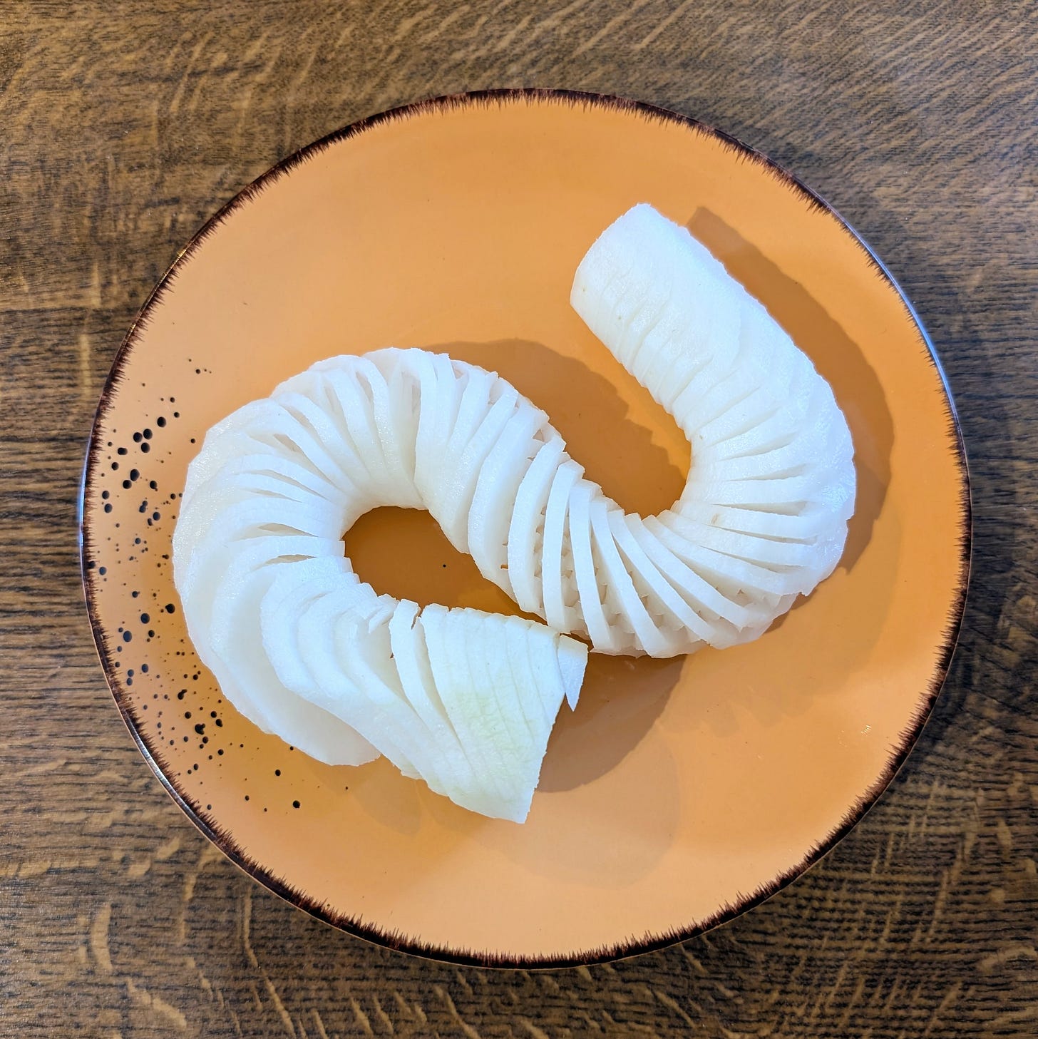A neatly spiralled length of white Bavarian radi (beer‑garden radish) sits on an orange plate with a dark rim. The thin, even slices curve in a wave-like shape, showing the radish’s crisp, translucent texture against the wooden surface beneath.