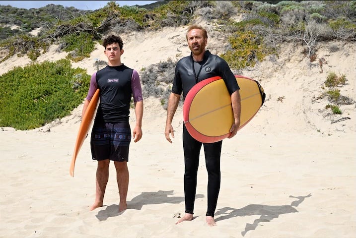 Nicolas Cage & Co-star on Beach Alt text: Scene from “The Surfer” (2024) showing Nicolas Cage and another surfer standing on a sandy beach, both holding boards and wearing wetsuits, looking off into the distance.