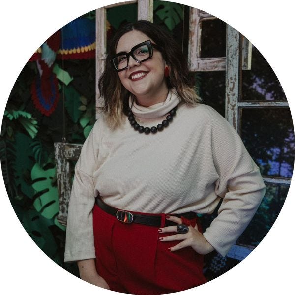 Foto da jornalista Renata Moniz usando uma calça vermelha e blusa de manga comprida bege. Ela usa óculos de grau de armação preta e tem a mão na cintura.
