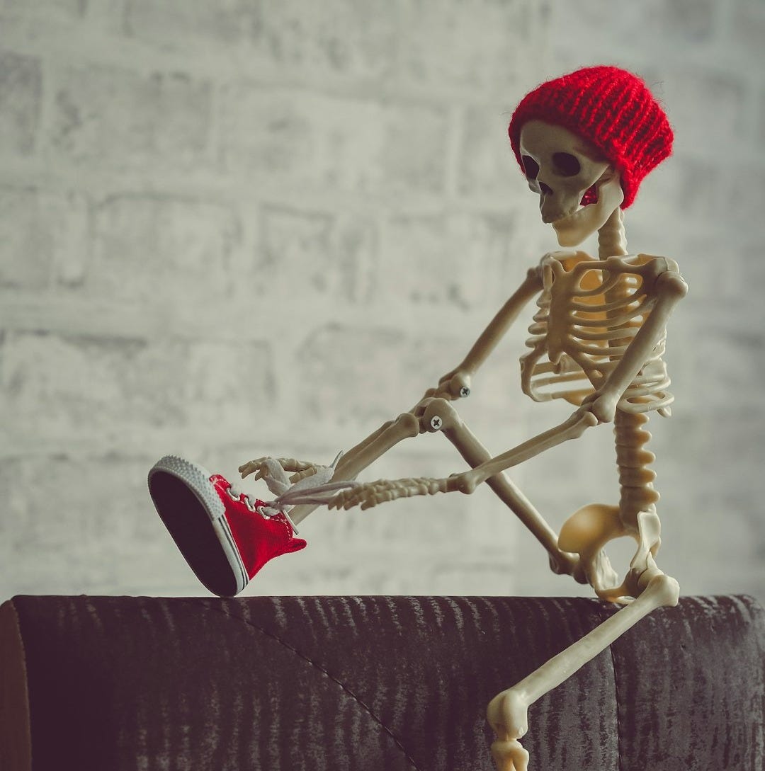 A skeleton figurine wearing a red beanie, lacing up red shoes on its feet A skeleton figurine wearing a red beanie, lacing up red shoes on its feet
