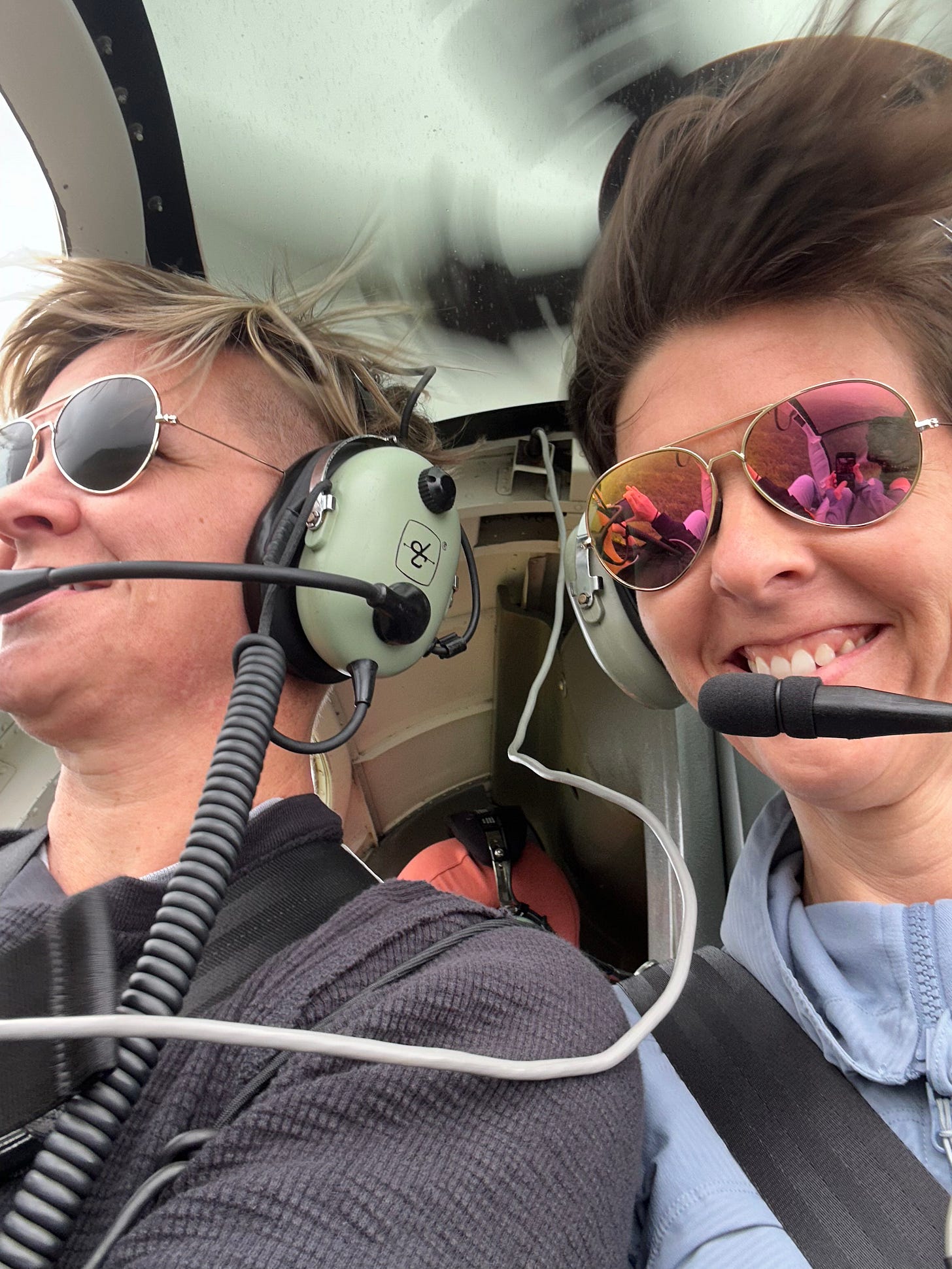 Two women wearing aviator sunglasses sit in a helicopter which is blowing their hair around.