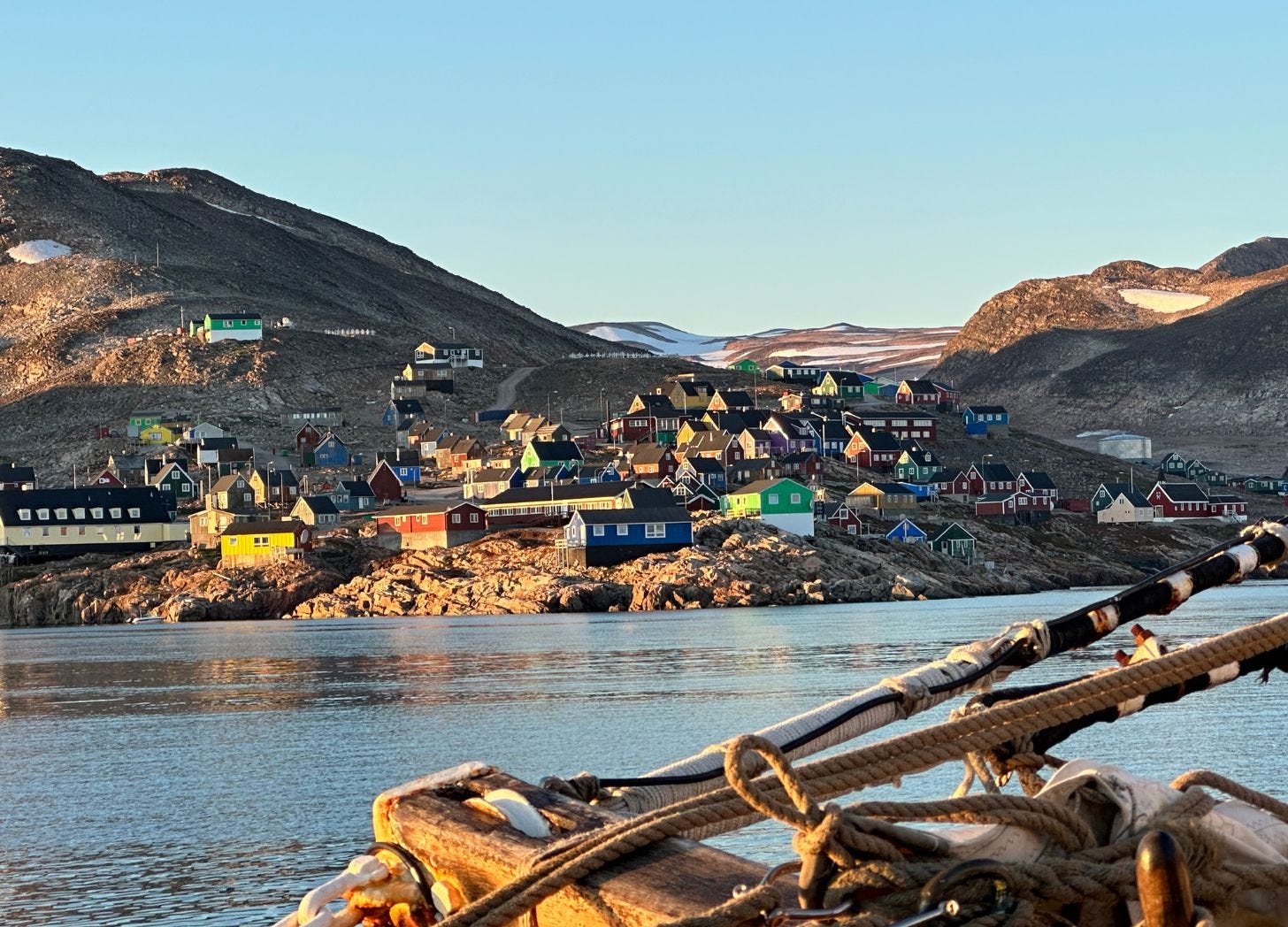 A view of Ittoqqortoormiit from the water at golden hour. Colored houses on rocky slopes are lit by evening sun. Wooden ship rigging and rope frame the lower right of the image. Mountains and open water in the middle distance.