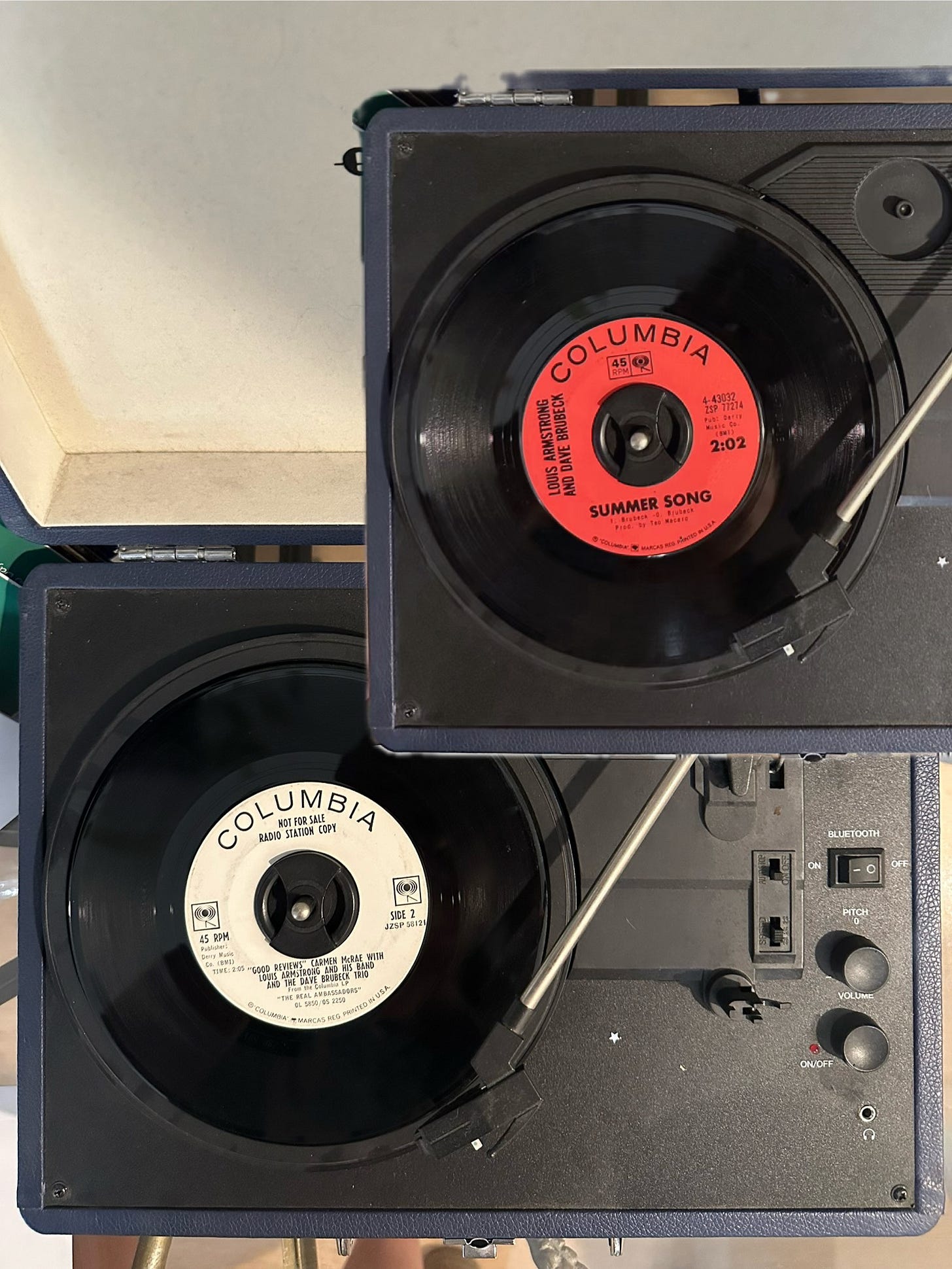 An overhead view of a dusty record player shows a 45 with a white label. At the top it says "Columbia, Not for Sale, Radio Station Copy." At the bottom it says "'Good Reviews' Carmen McRae with Louis Armstrong and His Band and the Dave Brubeck Trio, from the Columbia LP 'The Real Ambassadors.'" Superimposed in the top right is an image of another 45 on the same record player, this one with a red label. It reads "Columbia" across the top. On the left side it identifies the artists as Louis Armstrong and Dave Brubeck, and at the bottom it identifies the songwriters, I. Brubeck and D. Brubeck, and the producer, Teo Macero.