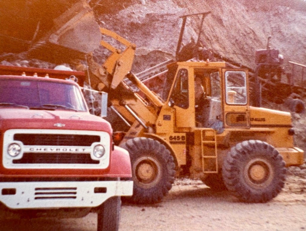 A red Chevrolet truck parked in front of a yellow loader machine on a construction site, showing a rugged landscape in the background. A red Chevrolet truck parked in front of a yellow loader machine on a construction site, showing a rugged landscape in the background.