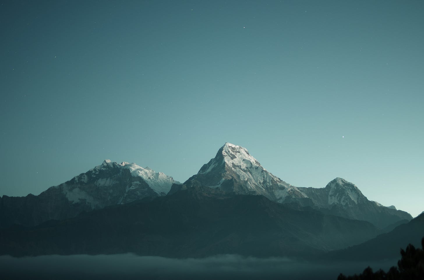 Snow-capped mountain peaks rising above a layer of fog under a deep teal sky.