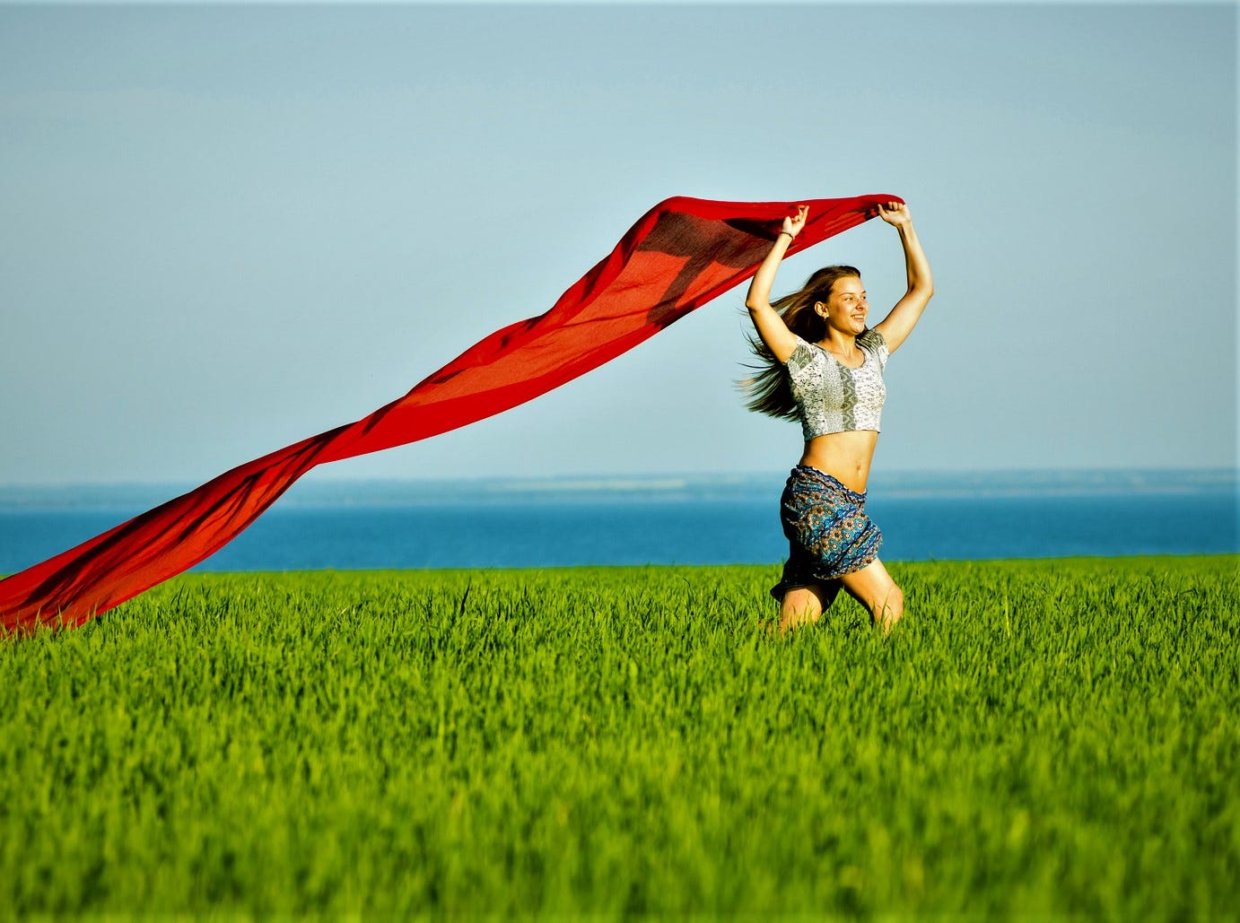 girl with long brown hair walking in field holding red piece of fabric