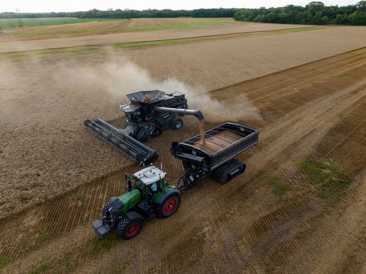 Aerial photograph of a Fendt combine unloading grain into a cart towed alongside by a Fendt tractor in a harvested field.