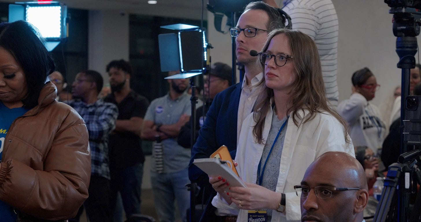 reporters watch, standing in front of lights holding a notebook
