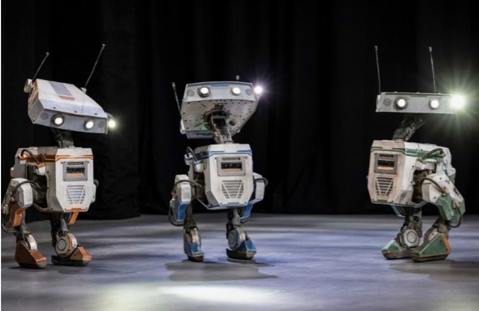 Three AI-powered Disney robots with a Star Wars-inspired design stand on a stage with a dark curtain backdrop. Each robot has a boxy, weathered body with mechanical limbs and a flat, rectangular head with glowing lights and antennas. These next-generation Disney theme park robots are part of an AI-driven collaboration with Nvidia and Google DeepMind, designed to move naturally and interact with guests using the Newton physics engine.