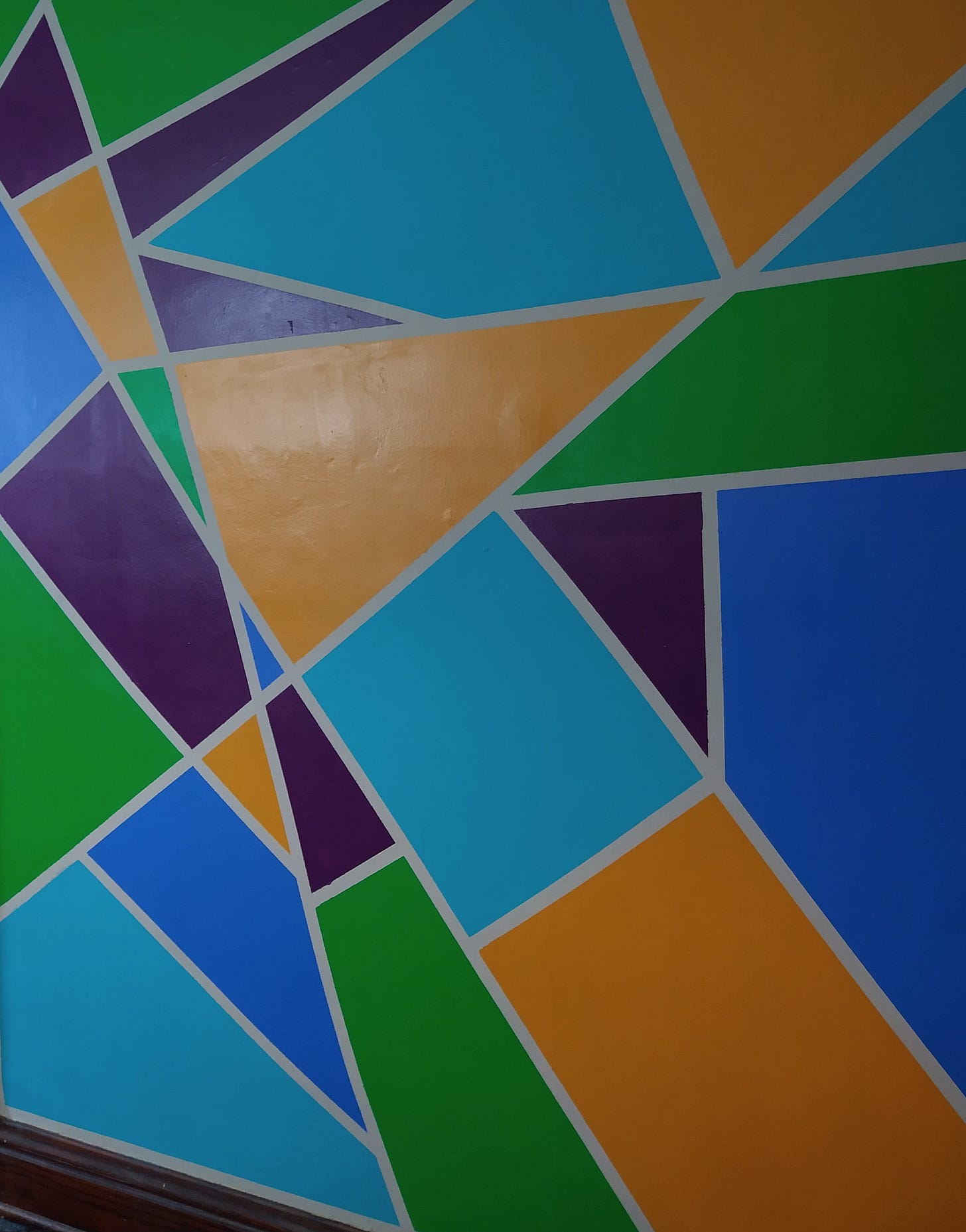A photograph of a wall painted yellow, green, teal, blue, and purple shapes with off-white outlines and a wooden baseboard. 