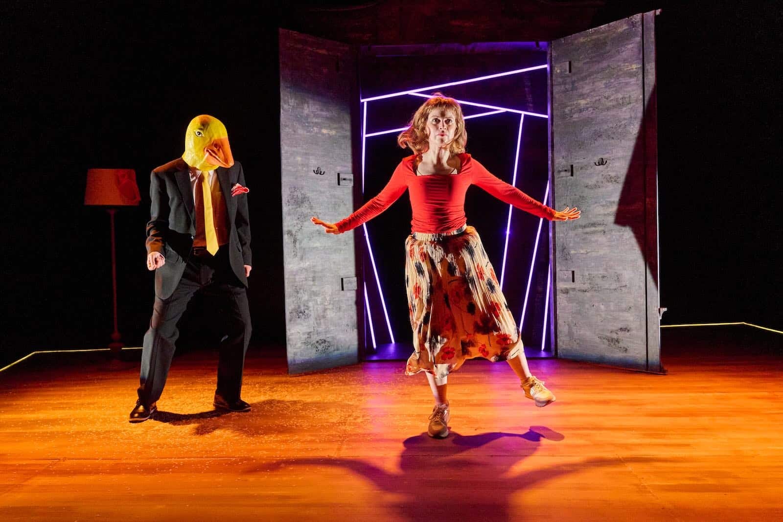 A man in a suit and a rubber-ducky mask looks on as a woman dances frantically in front of a large wardrobe with its doors open