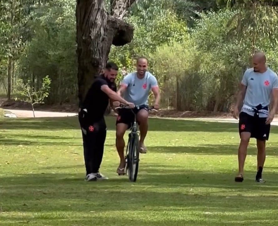 serginho em um campo, desequilibrado sobre a bicicleta, um homem o ajuda a se equilibrar, maicon observa à distância com um sorriso pueril