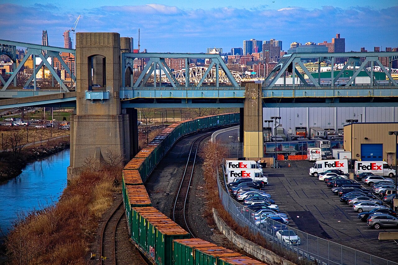 An aerial view of a rail line curving beneath a steel truss bridge with a FedEx distribution facility and parked vehicles below, set against a city skyline with multiple bridges spanning a waterway.