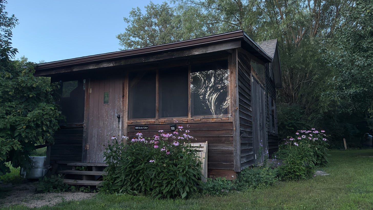 A small, wooden cabin with purple cone flowers in front. A small black sign reads. “1880 Settler’s Cabin.”