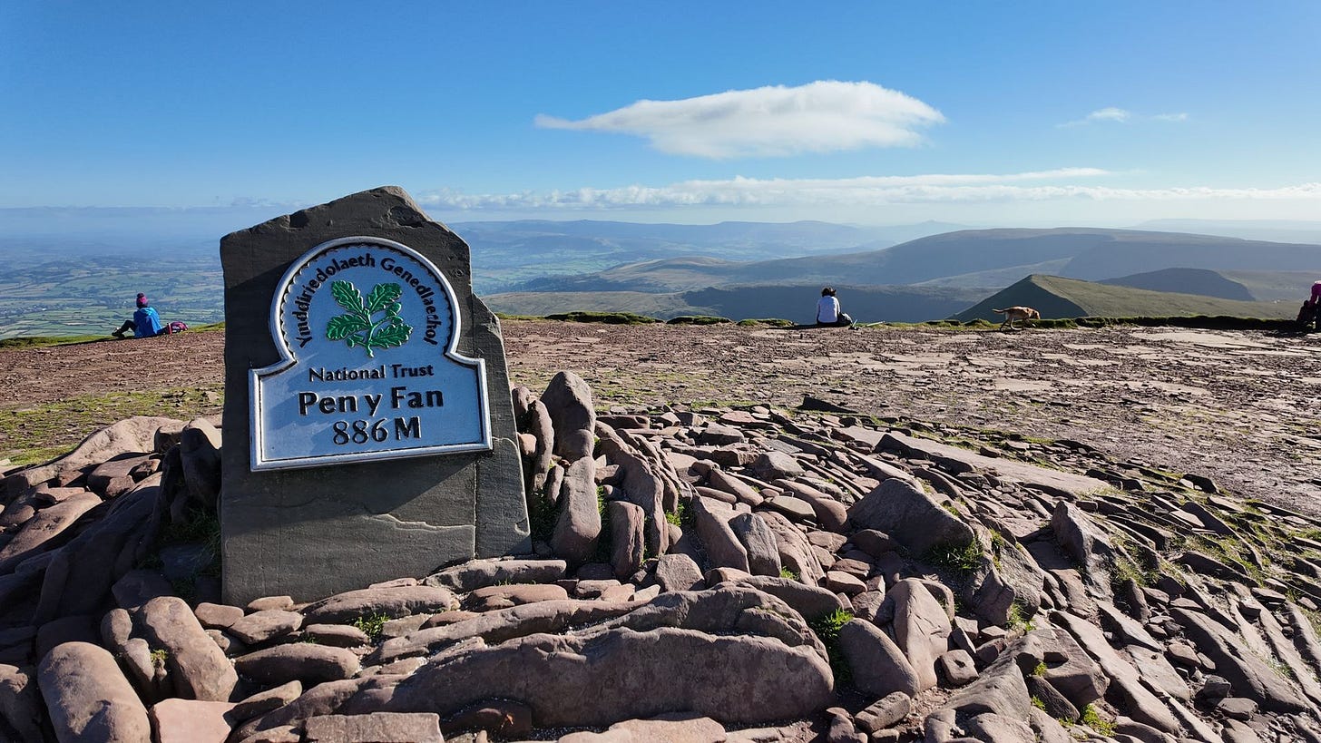 A stone summit marker bearing a National Trust plaque that reads "Pen y Fan 886 M" stands prominently amidst loose reddish-brown rocks. In the background, hikers sit on the broad, flat mountain plateau, taking in sweeping views of the rolling green valleys and distant hills under a bright blue sky.
