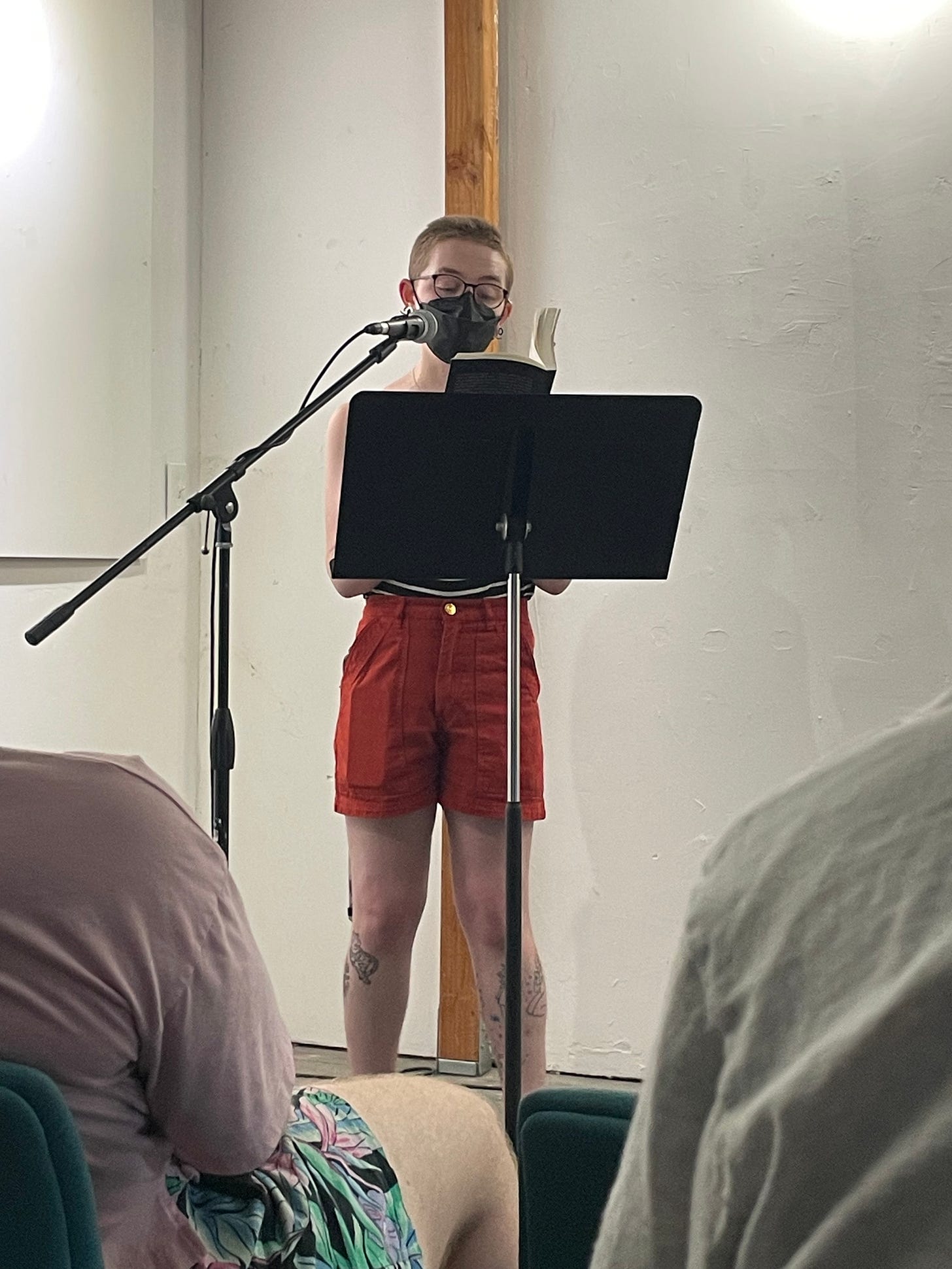 Cavar, wearing a black and white striped tank top and red shorts, stands behind a music stand and reads from Failure to Comply.