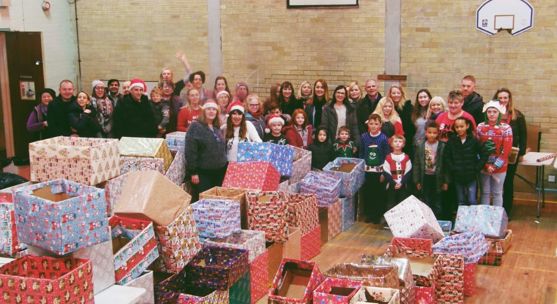 A crowd of people smiling into the camera in a gymnasium, with a pile of wrapped boxes in front of them A crowd of people smiling into the camera in a gymnasium, with a pile of wrapped boxes in front of them
