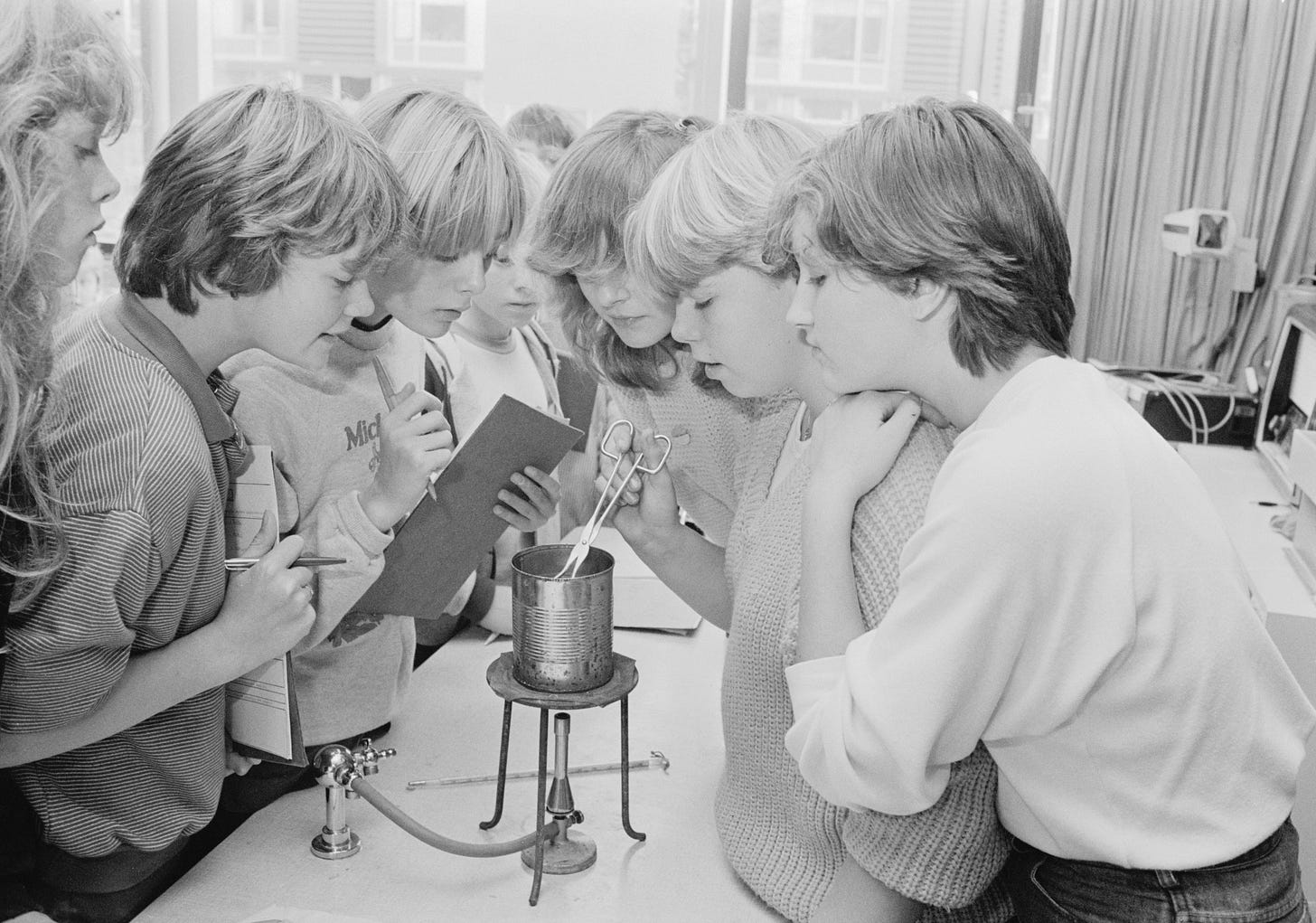 A group of 7 middle school kids stand around a lab bench conducting an experiment.