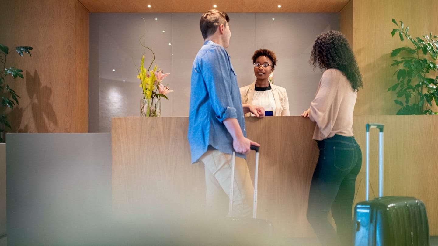 A couple with luggage talks to a female receptionist at a hotel front desk. A couple with luggage talks to a female receptionist at a hotel front desk.