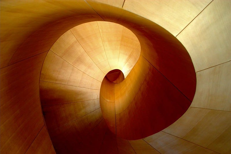 Photograph looking upward into a spiraling architectural interior made of warm, honey-toned wood panels. Smooth curved surfaces sweep inward in a continuous spiral toward a small circular opening at the center, where soft light enters. The effect is organic and enveloping, like being inside a shell or a living form. Photograph looking upward into a spiraling architectural interior made of warm, honey-toned wood panels. Smooth curved surfaces sweep inward in a continuous spiral toward a small circular opening at the center, where soft light enters. The effect is organic and enveloping, like being inside a shell or a living form.