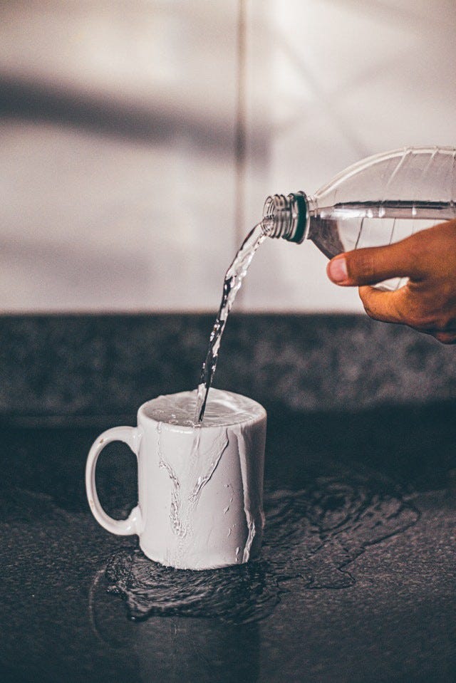 Water being poured from a bottle into a mug already overflowing with water. The additional water is spilling beneath it. Water being poured from a bottle into a mug already overflowing with water. The additional water is spilling beneath it.