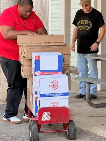 A person in a red shirt loads a red hand truck stacked with cardboard boxes, including several large Coffee mate French Vanilla cartons. Nearby, another person wearing a black T‑shirt with a yellow Batman logo and a camo cap stands beside a gray picnic table. The scene is an outdoor loading or side area next to a building with siding, columns, and concrete pavement.