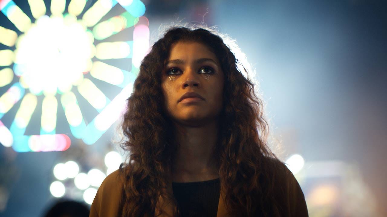 A cinematic close-up of Zendaya as Rue Bennett in Euphoria, featuring her signature glitter "tears" makeup under the eyes, set against the blurred, glowing lights of a carnival Ferris wheel at night.