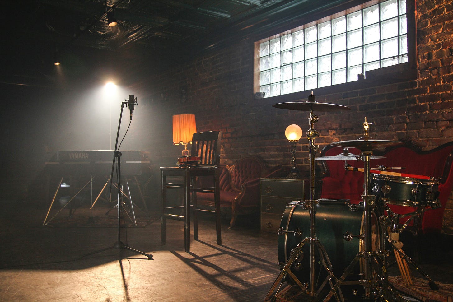 photo of a dark room with a spot light. The walls are brick and there's a small window. Several empty chairs and instruments form a semi-circle around the microphone. It's a scene where you can imagine hearing great local music.