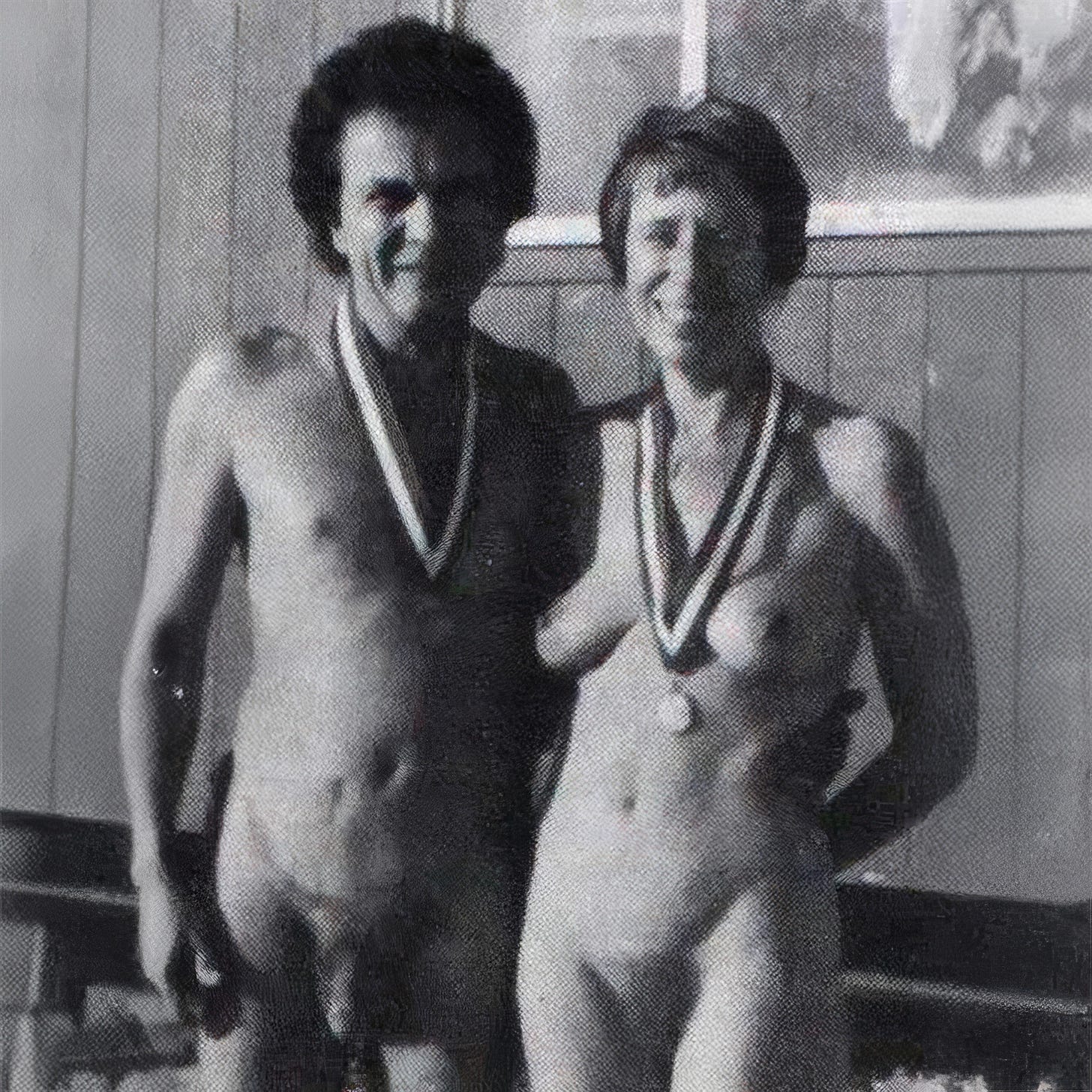 Black-and-white archival photo of two nude men standing side by side indoors, smiling and wearing medals on ribbons around their necks, with one man’s arm around the other at what appears to be an awards or festival setting.