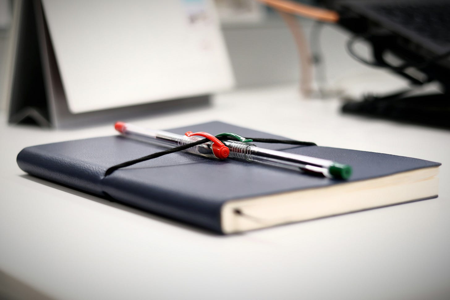 A close-up of a black closed journal resting on a desk, with two pens and a black elastic loop secured across it. A close-up of a black closed journal resting on a desk, with two pens and a black elastic loop secured across it.
