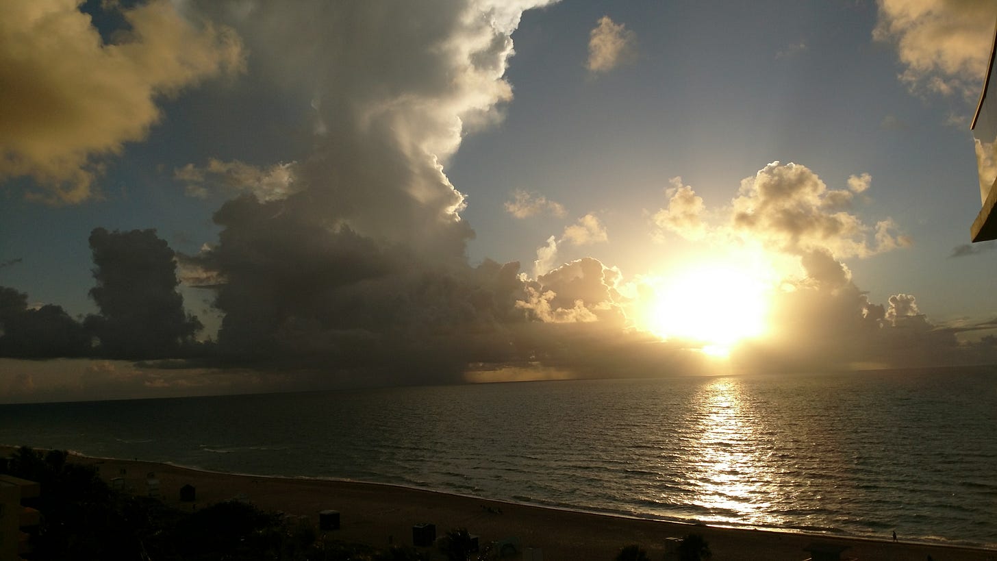 Sunrise over the ocean with golden light breaking through dramatic clouds, reflecting across calm waves and illuminating the shoreline below.