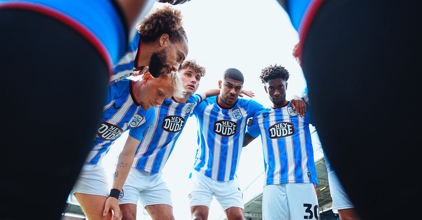 Huddersfield Town players form a huddle, with the Accu Stadium in the background. 