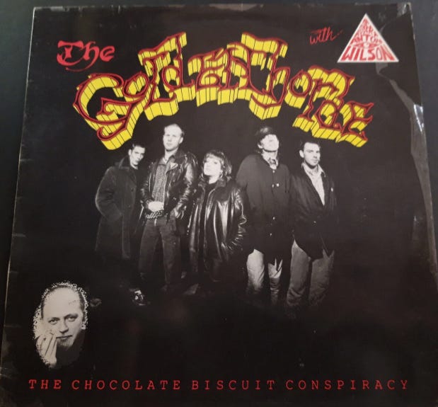 An album cover from the band called The Golden Horde. It features a photo of the band members. Beneath is the album title of The Chocolate Biscuit Conspiracy. In the upper right is an eye in the triangle with the name Robert Anton Wilson within. In the bottom left corner is a headshot of Robert Anton Wilson. The whole image looks like it was stolen from an ebay listing. Fnord. But what do I know.
