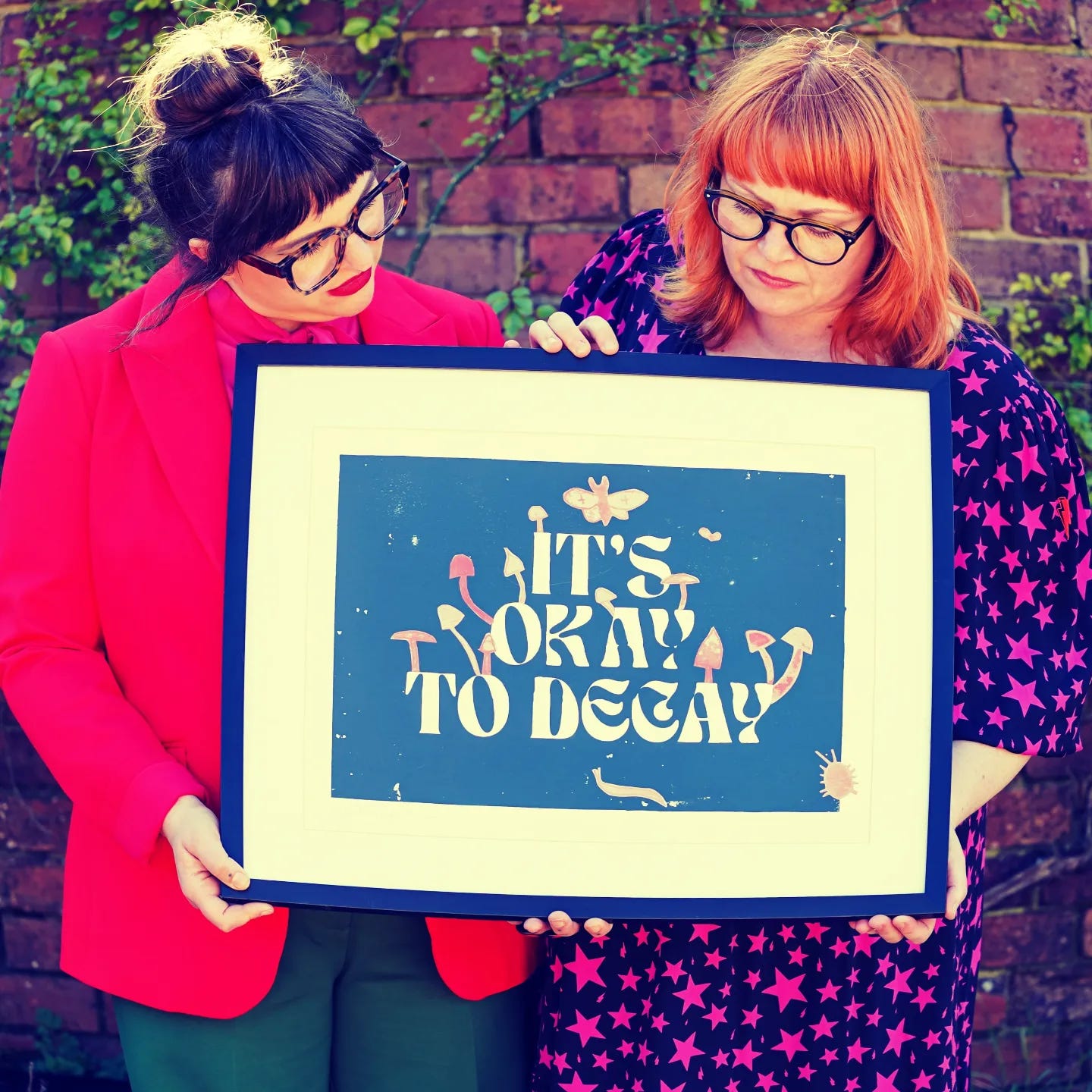 Two white women wearing glasses holding a framed print that says IT'S OKAY TO DECAY. Two white women wearing glasses holding a framed print that says IT'S OKAY TO DECAY.
