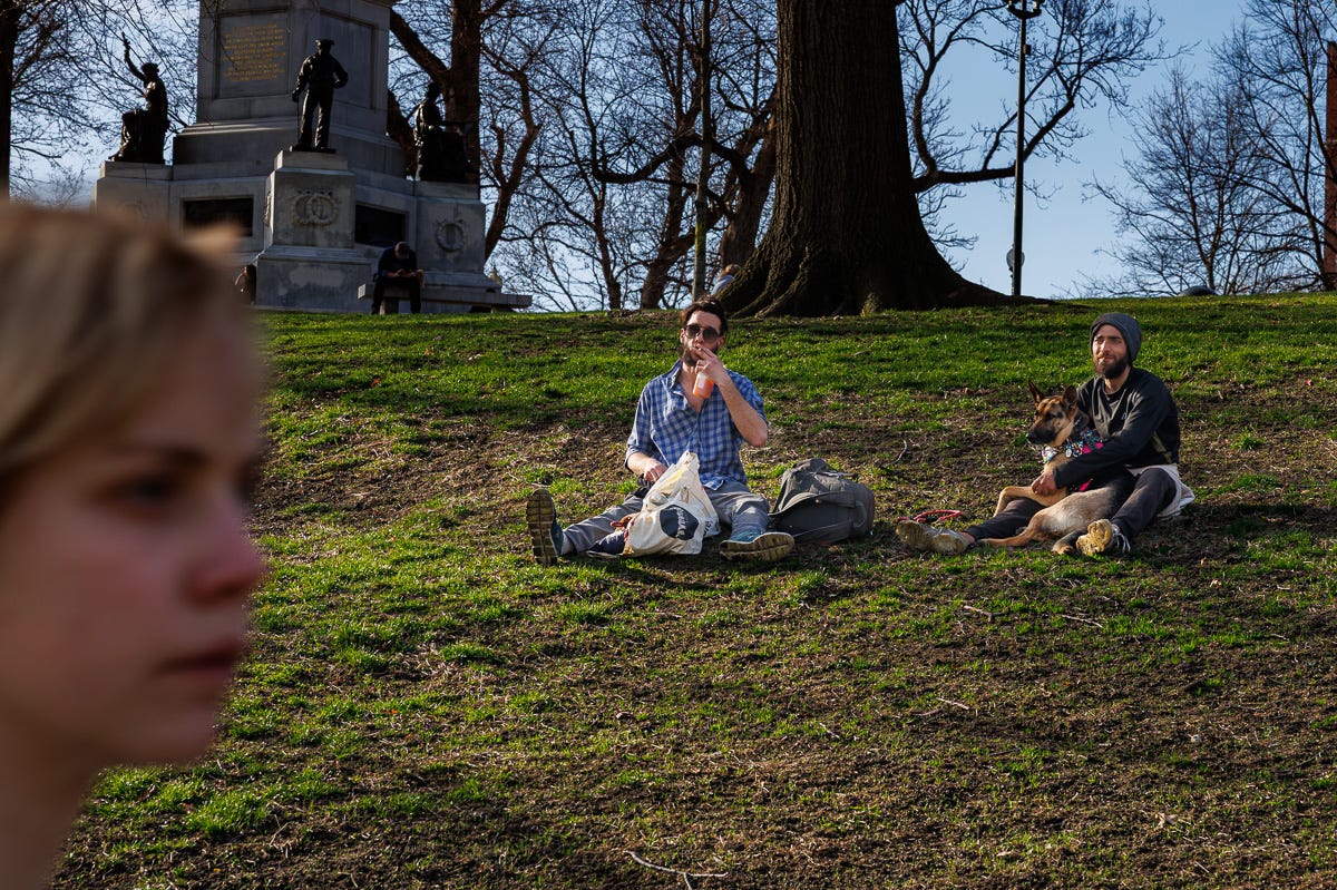 Two men sitting on a grassy hill at Boston Common, one eating and the other with a dog, while a blurry face passes in the foreground and a statue looms in back. Two men sitting on a grassy hill at Boston Common, one eating and the other with a dog, while a blurry face passes in the foreground and a statue looms in back.