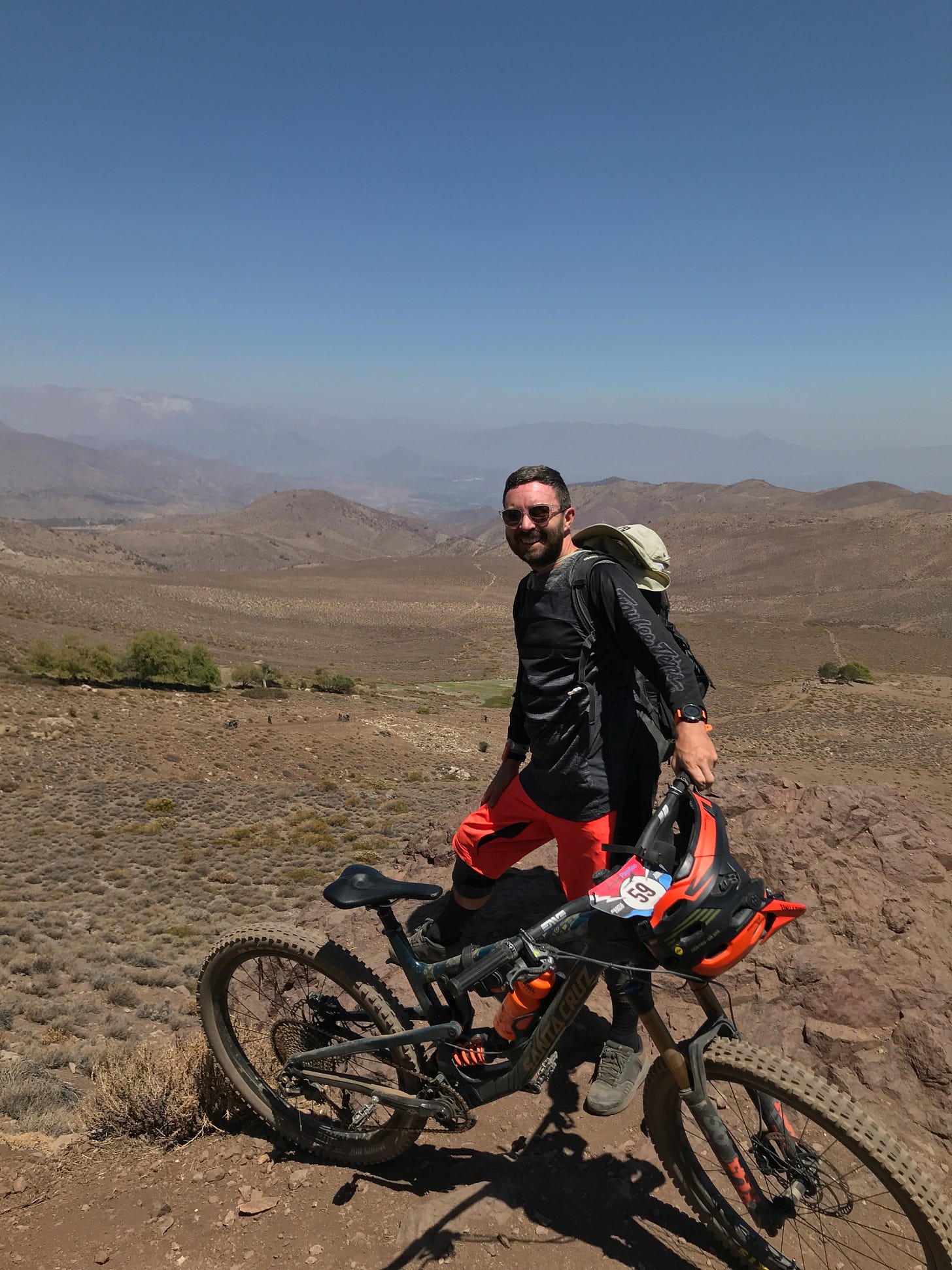 Richard Duke standing with his mountain bike on a high desert ridge in Chile, wearing sunglasses and cycling gear with sprawling mountain valleys behind him.