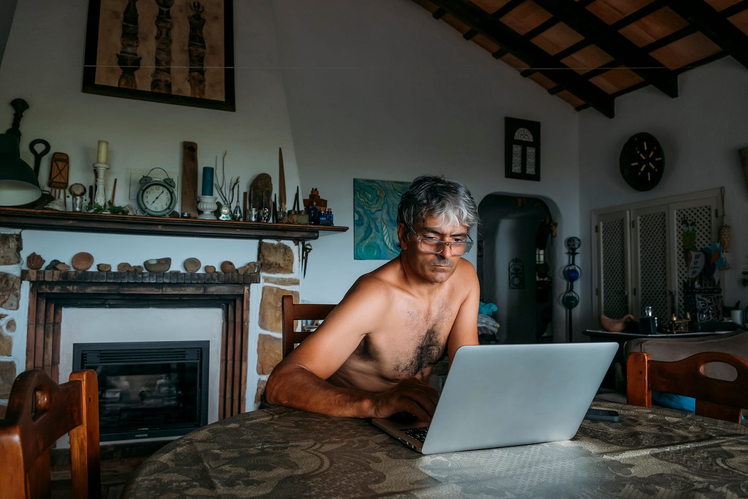 A man works on a laptop at his kitchen table, undressed at home.