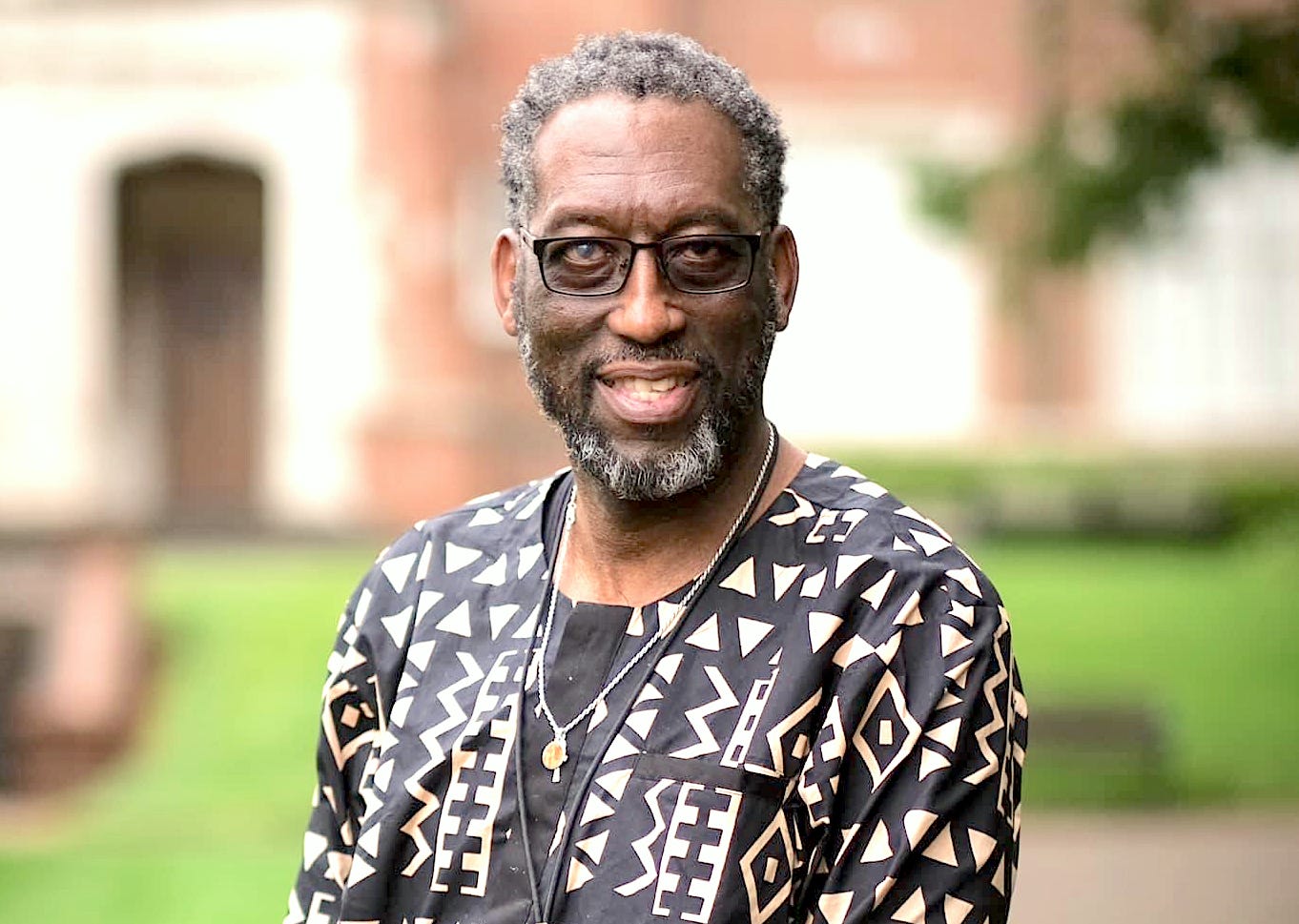 Kamal faces the camera. He wears glasses, has short salt-and-pepper grey hair and beard, and wears a shirt with an Afrocentric pattern in black and white.