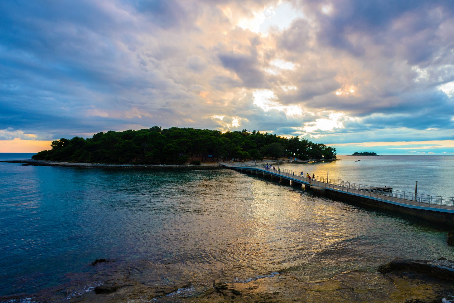 The footbridge connecting Koversada's naturist island to the mainland at Vrsar, stretching across calm water at sunset with a wooded shoreline and a small offshore islet in the distance. The footbridge connecting Koversada's naturist island to the mainland at Vrsar, stretching across calm water at sunset with a wooded shoreline and a small offshore islet in the distance.