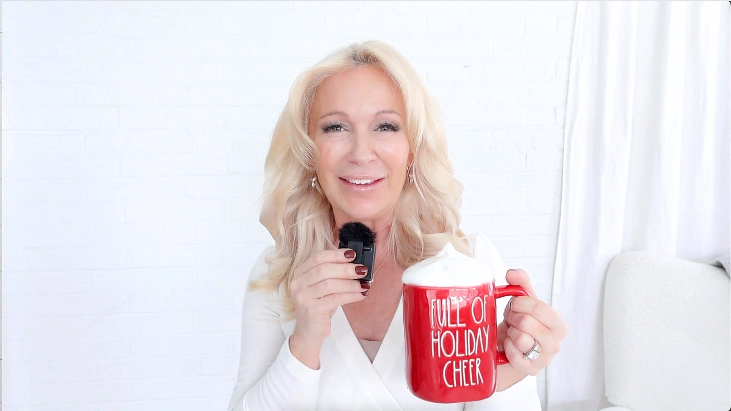 A woman smiling and holding a festive red mug that says 'FULL OF HOLIDAY CHEER'. She has long blonde hair and is wearing a white outfit against a light-colored background.