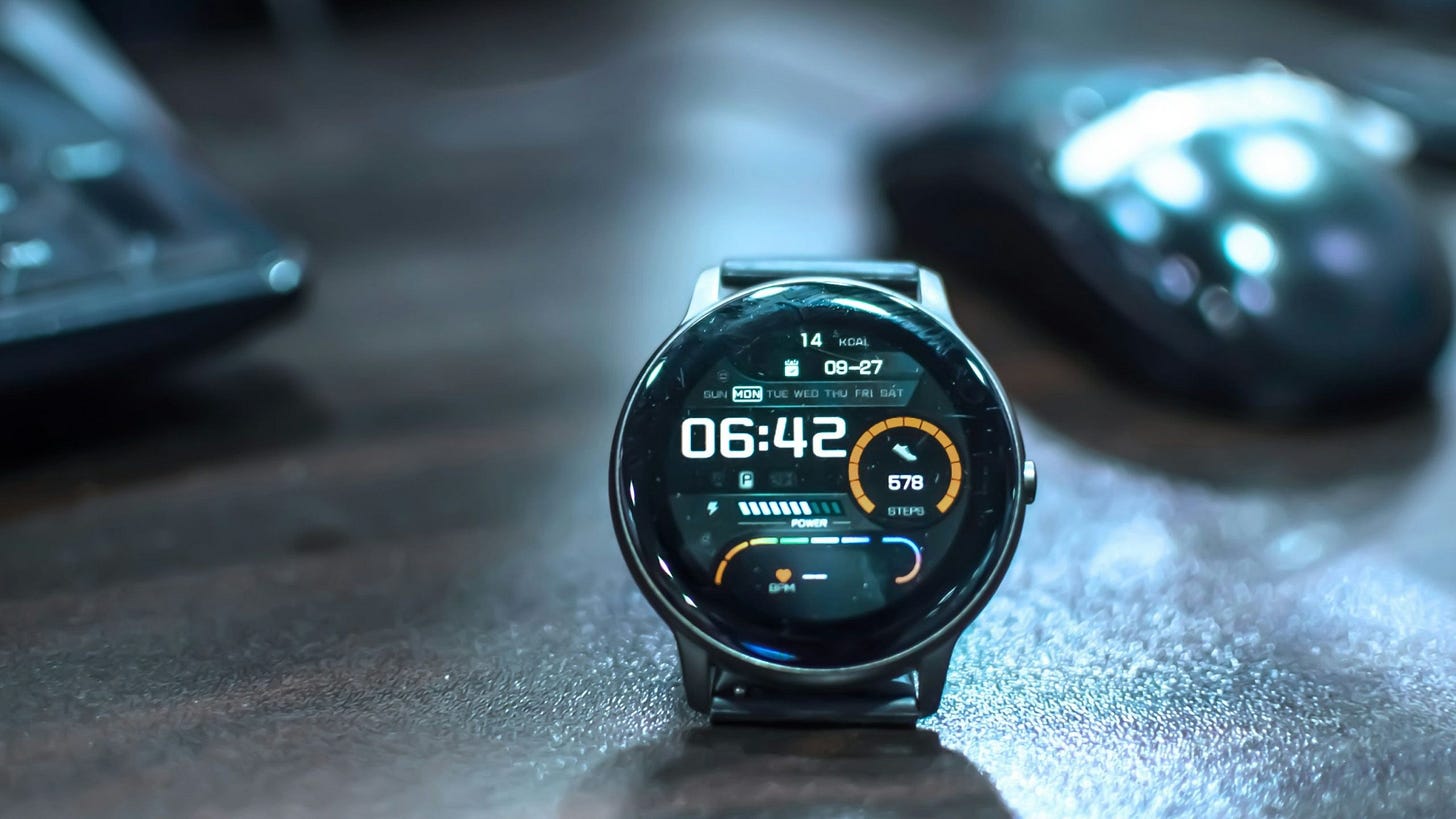 Close-up of a black smartwatch displaying the time 06:42, date 09-27, day Monday, steps count 578, and power status, placed on a textured surface with a blurred keyboard and mouse in the background.