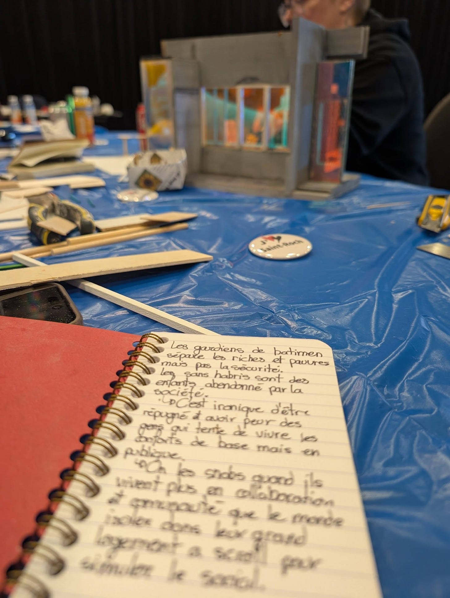 A spiral notebook open on a blue plastic-covered table, covered in handwritten French text. In the background, a detailed miniature building constructed from cardboard and mixed materials, and a J'aime St-Roch button. A person in a dark jacket and glasses is partially visible in the background.