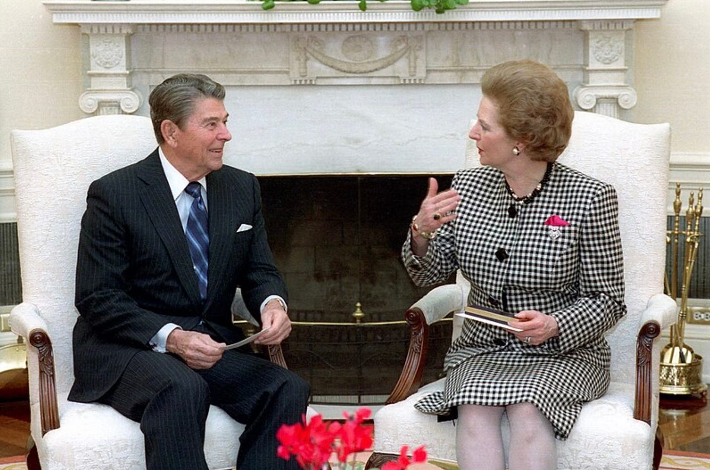 Reagan and Thatcher look like they are having a lively discussion; Reagan smiles and listens as Thatcher has her hands in the air as if she is making a point. the two are seated on white chairs, in front of a carved fireplace. Reagan and Thatcher look like they are having a lively discussion; Reagan smiles and listens as Thatcher has her hands in the air as if she is making a point. the two are seated on white chairs, in front of a carved fireplace.
