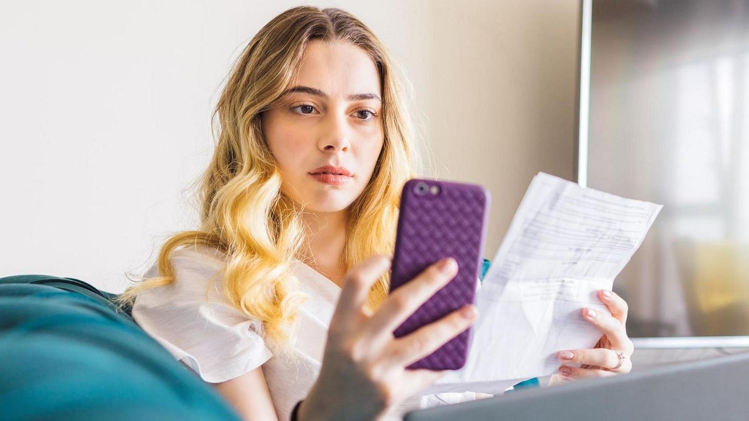 A young blonde woman with a look of concentration stares at her smartphone while holding a bill in her other hand A young blonde woman with a look of concentration stares at her smartphone while holding a bill in her other hand