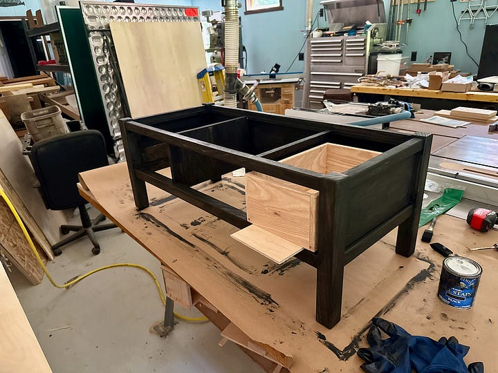 Two workshop views of a wooden bench project. Left: unfinished bench frame clamped together on a workbench. Right: the same bench painted dark, with a drawer box slid partway into place.