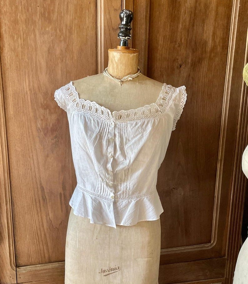 May include: A white, vintage-style blouse with a square neckline and delicate lace trim. The blouse features small buttons down the front and a ruffled hem. The garment is displayed on a vintage dress form.