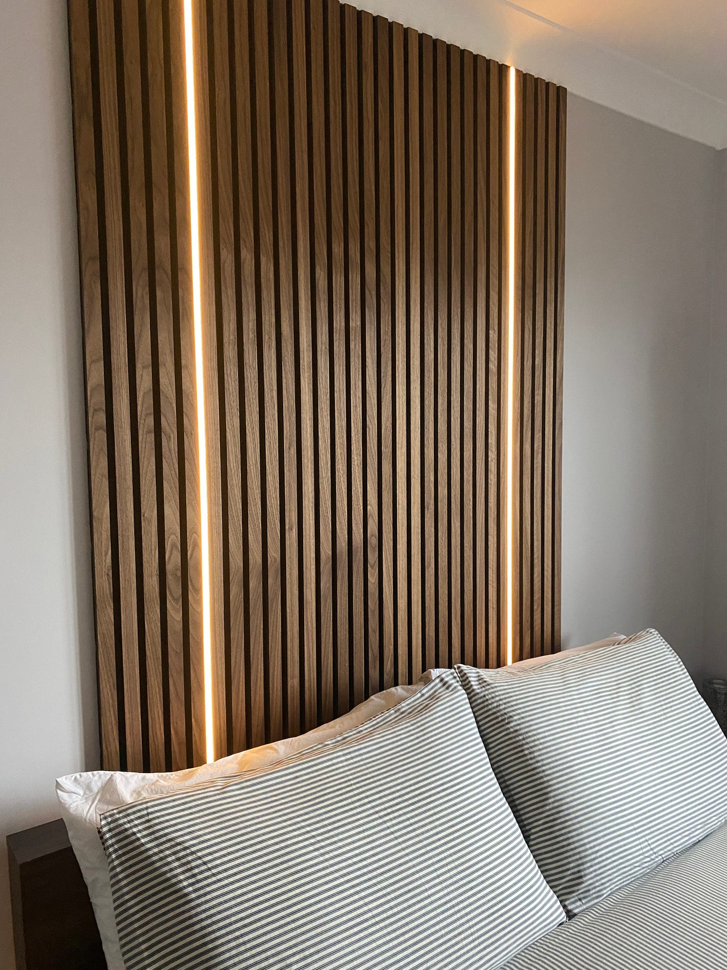 bedroom with dark walnut wood panelling applied over the bed and two LED strip lights positioned within the panelling