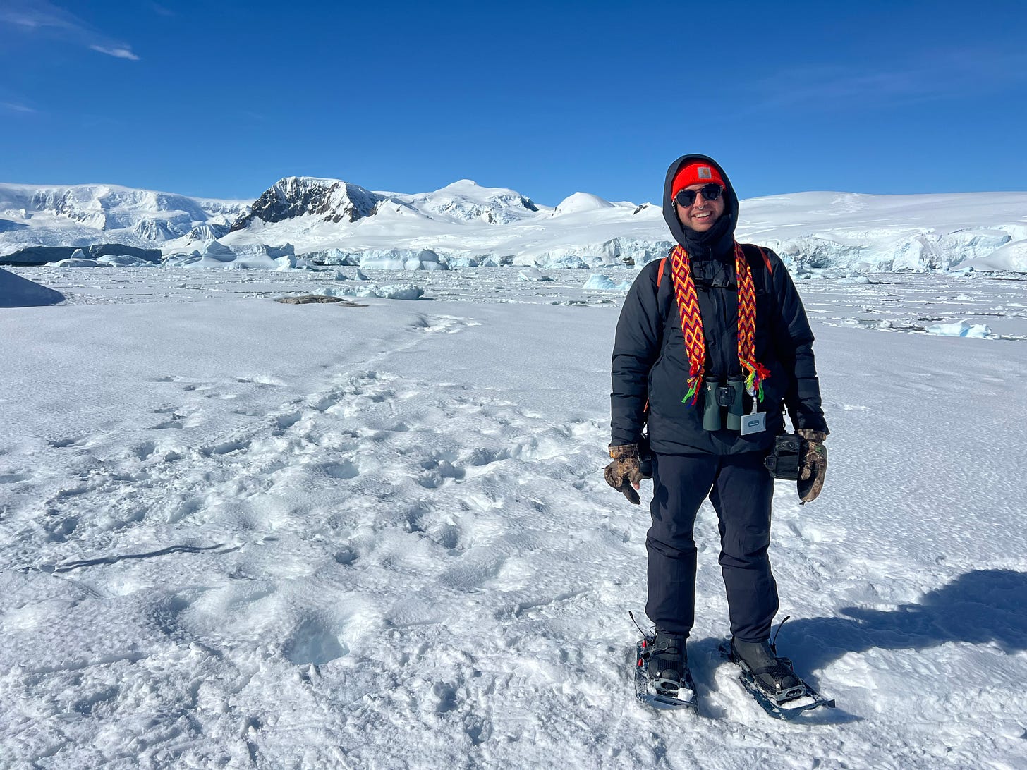 a person in a down jacket, snow pants, and snowshoes standing on the snow and ice on a sunny day with glaciated mountains and ice-covered water in the background