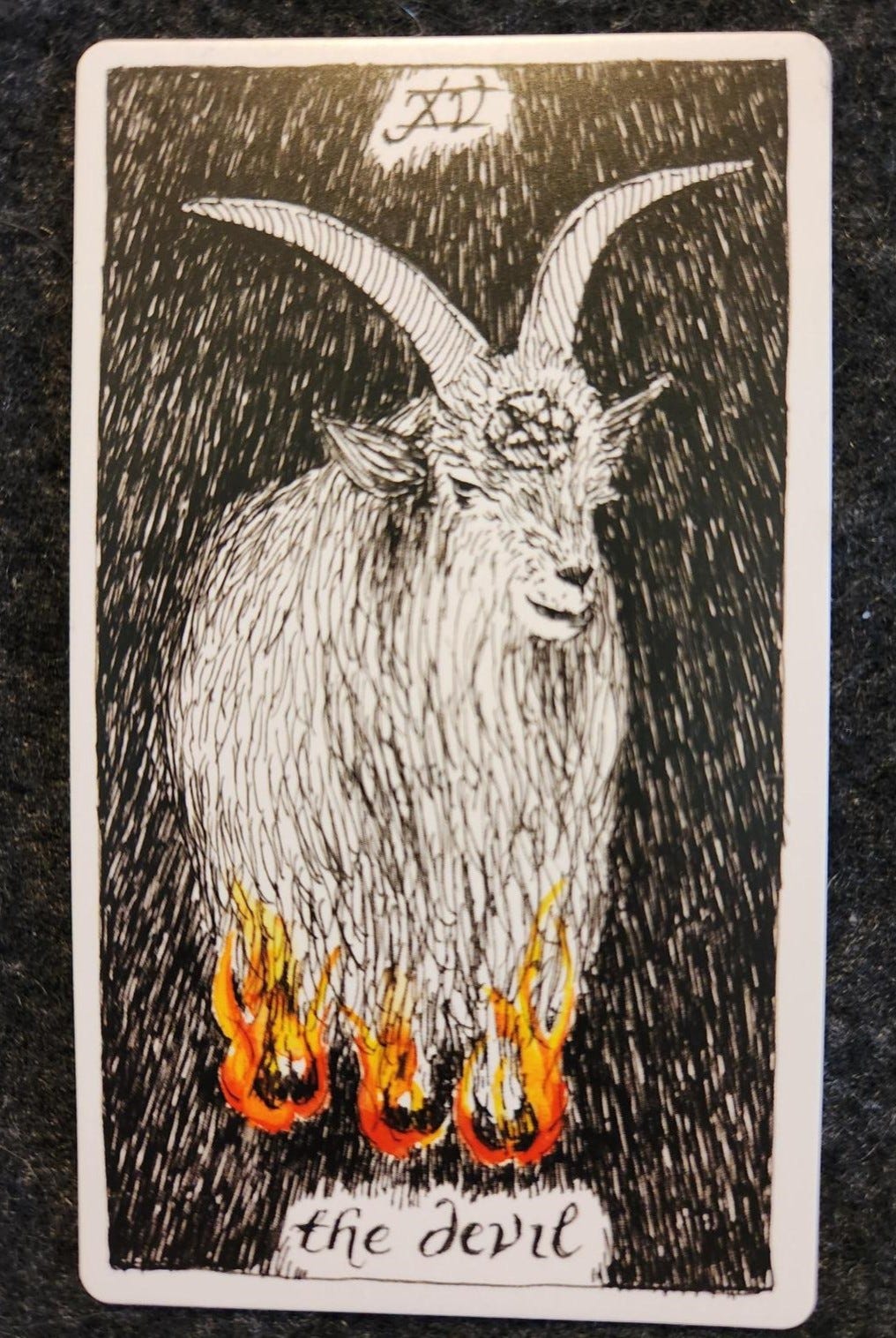A tarot card labelled XV the devil. A goat with long horns. Its hooves are encased in red, orange, and yellow flames. Otherwise, the card is fully black and white. On its head is an inverted pentacle.