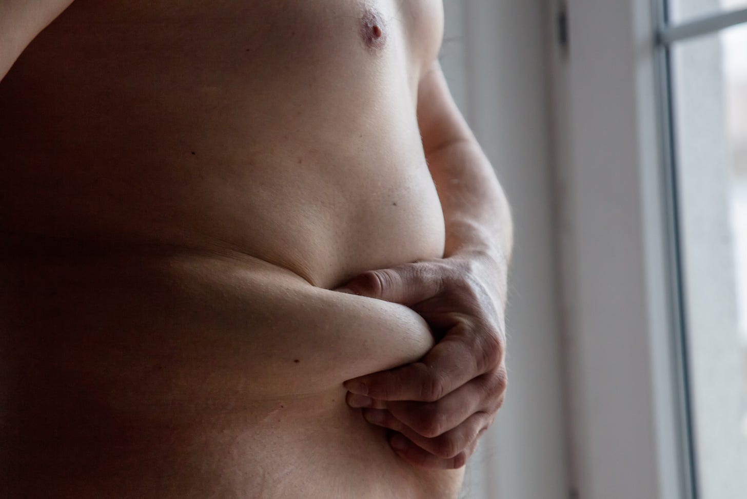 Close-up of a nude torso near a window, with one hand gently holding a fold of the abdomen in soft natural light.